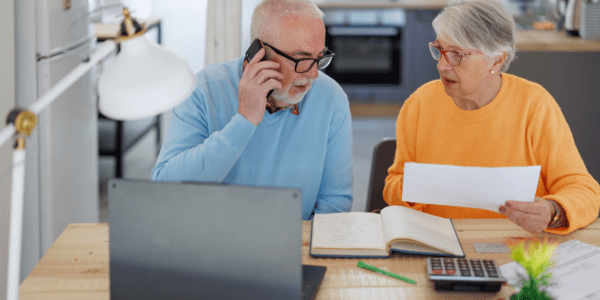 Een ouder echtpaar zit aan een tafel met een laptop, papieren, een notitieboek en een rekenmachine. De man belt terwijl de vrouw een document vasthoudt en informatie bespreekt.